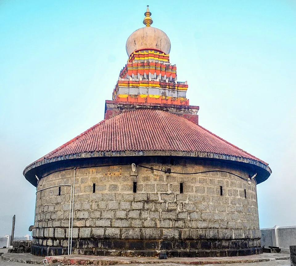 The round stone shrine of the Karinjeshwara temple with a red roof and a tiered coloured tower, against a blue sky