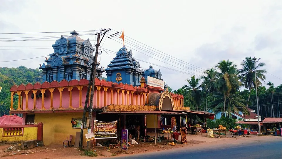The blue-grey roofs of the Kudupu Ananthapadmanabha temple seen from the road, above a pink arcade and small shops, with a dark hill behind