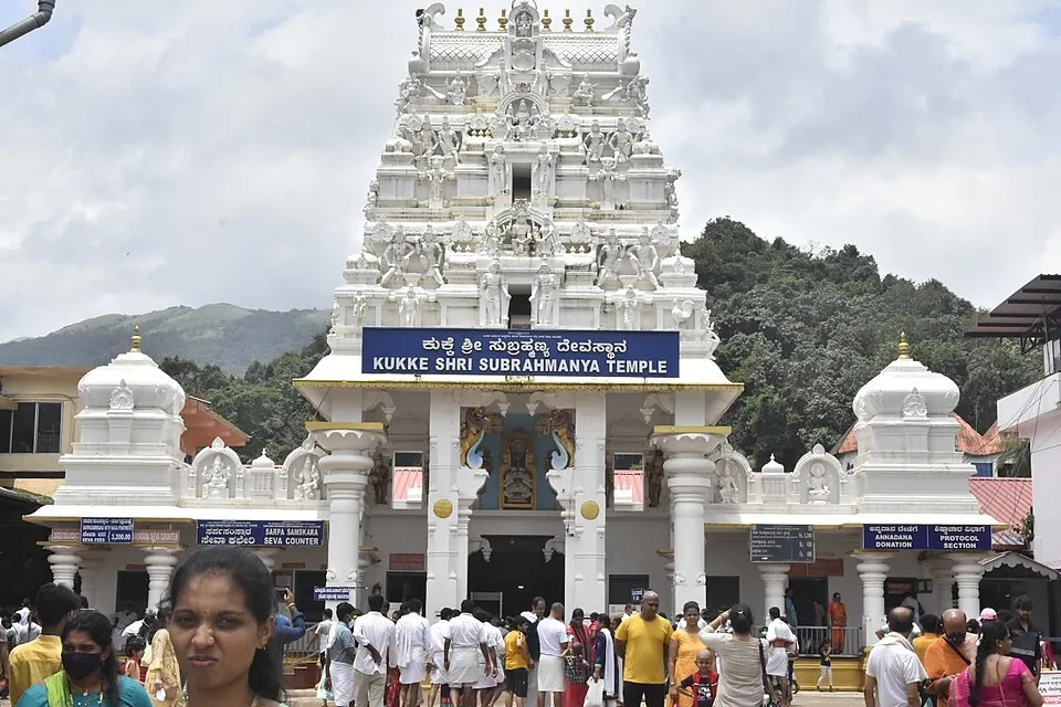 The white gateway tower of the Kukke Subramanya temple with its name board, the entrance building and a crowd of visitors, with forested hills behind