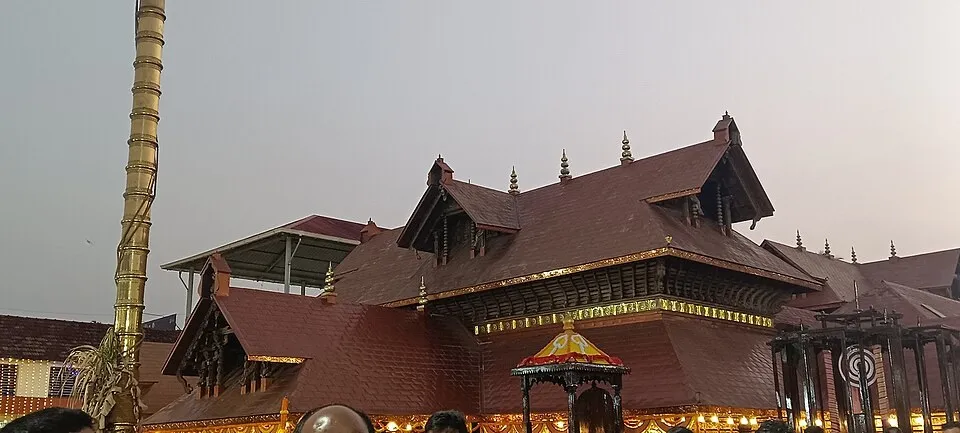 The tiered tiled roofs of the Polali Rajarajeshwari temple beside a tall brass flagpole, seen under a grey evening sky