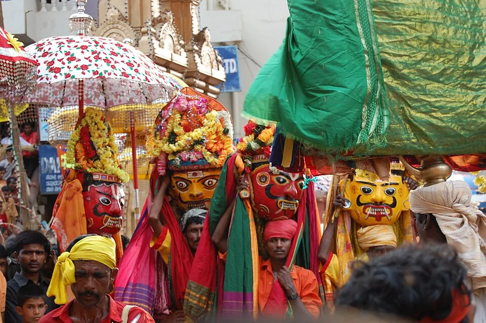 Large painted mask-faced dolls carried on men's heads in a procession, under flowered umbrellas