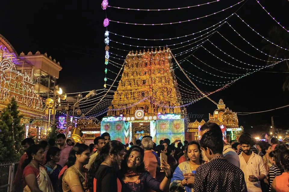 The lit gopuram of the Kudroli temple at night under strings of lights, with a crowd of people in the foreground
