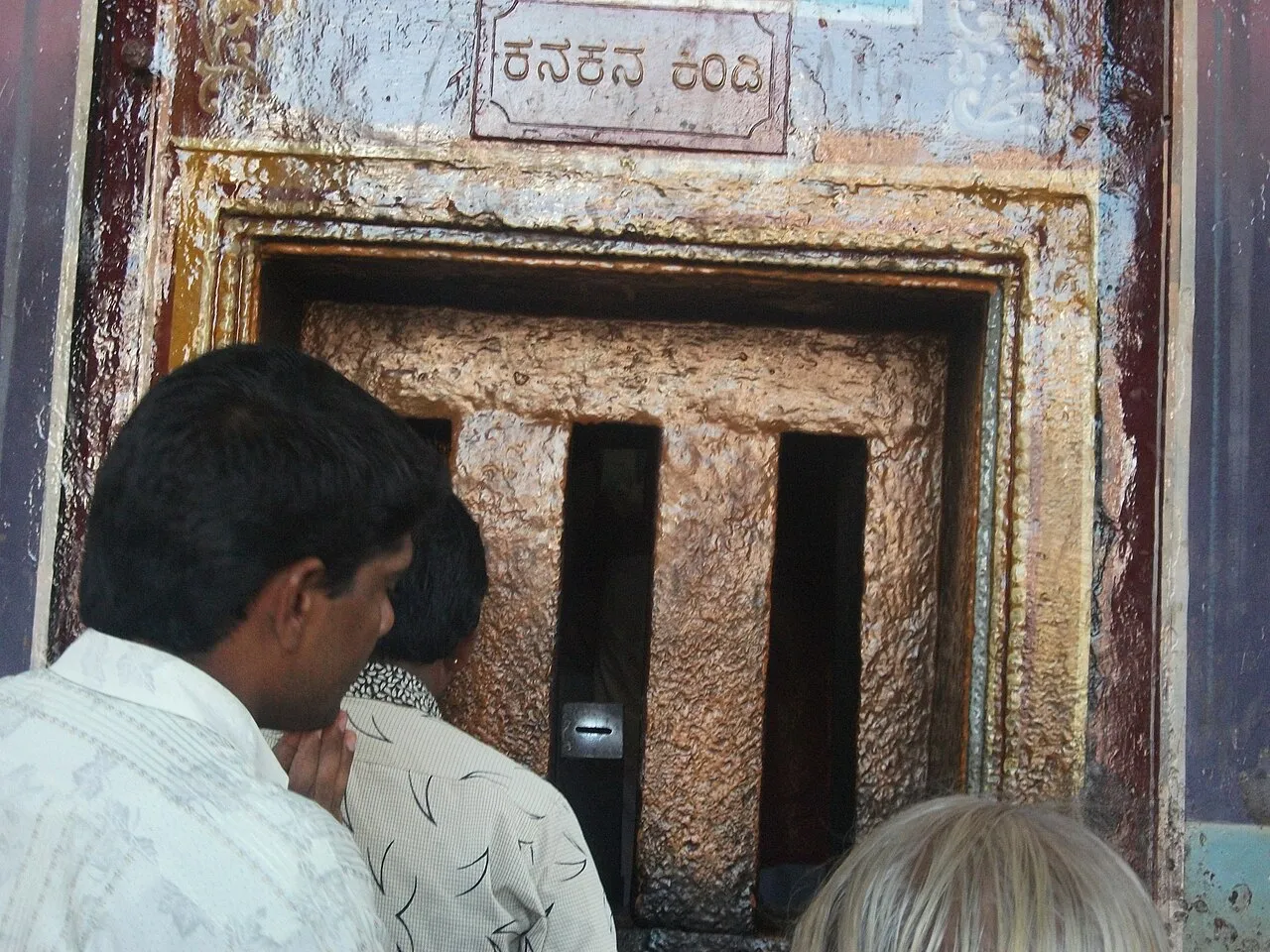 Pilgrims peering through Kanakana Kindi, a small barred window in an old stone wall with its name painted above