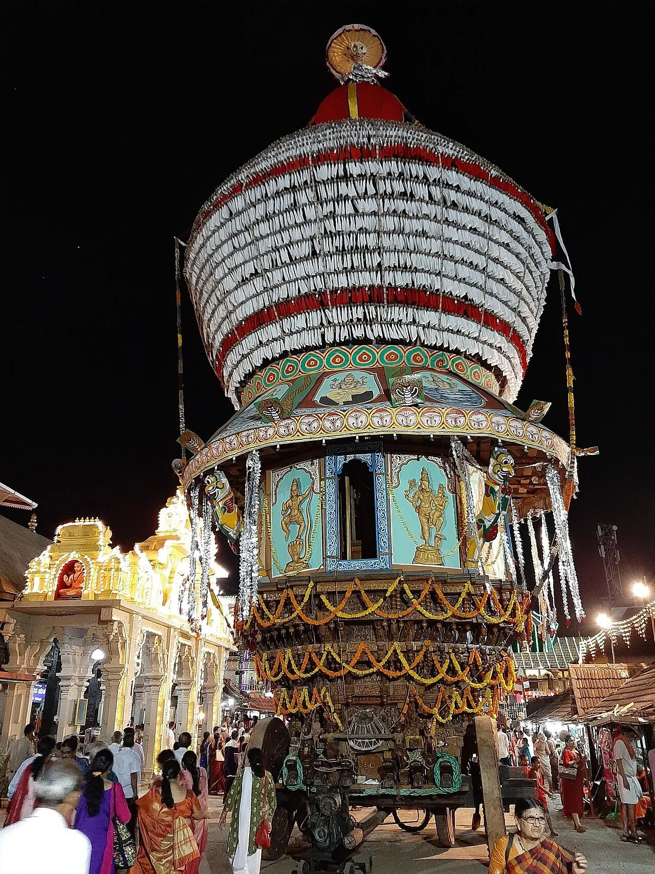 A towering wooden temple chariot, decked with cloth and lights, at night during the Paryaya festival