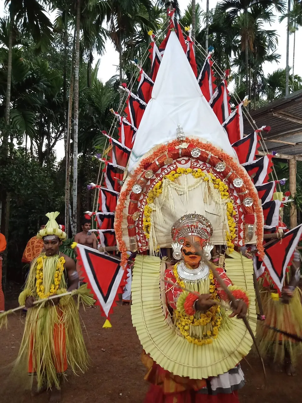 A performer as one of the Ullakulu, holding a bow and arrow, his face painted orange under a silver crown, before a towering frame of white cloth, flowers and red and black flags