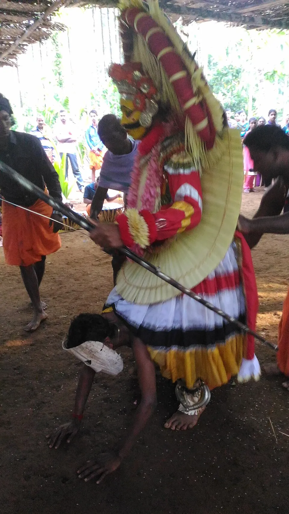 A performer as the Ullakulu daiva, in a tall red and gold headdress and a fan of tender palm leaves, holds a long bow while a man bends to the ground at his feet