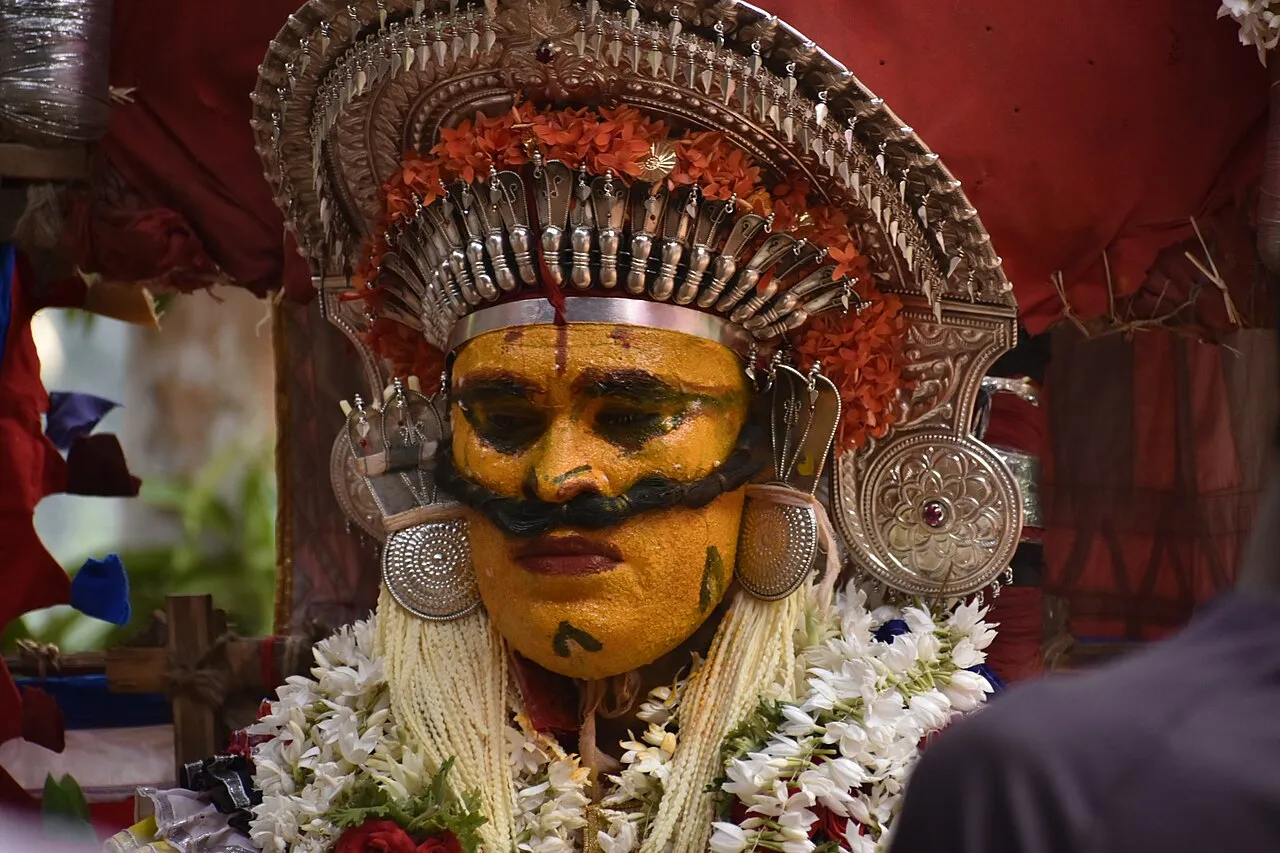 Close-up of a performer as an Ullakulu daiva, face painted yellow with a thick black moustache, under a tall silver crown with silver ear discs, garlanded with jasmine